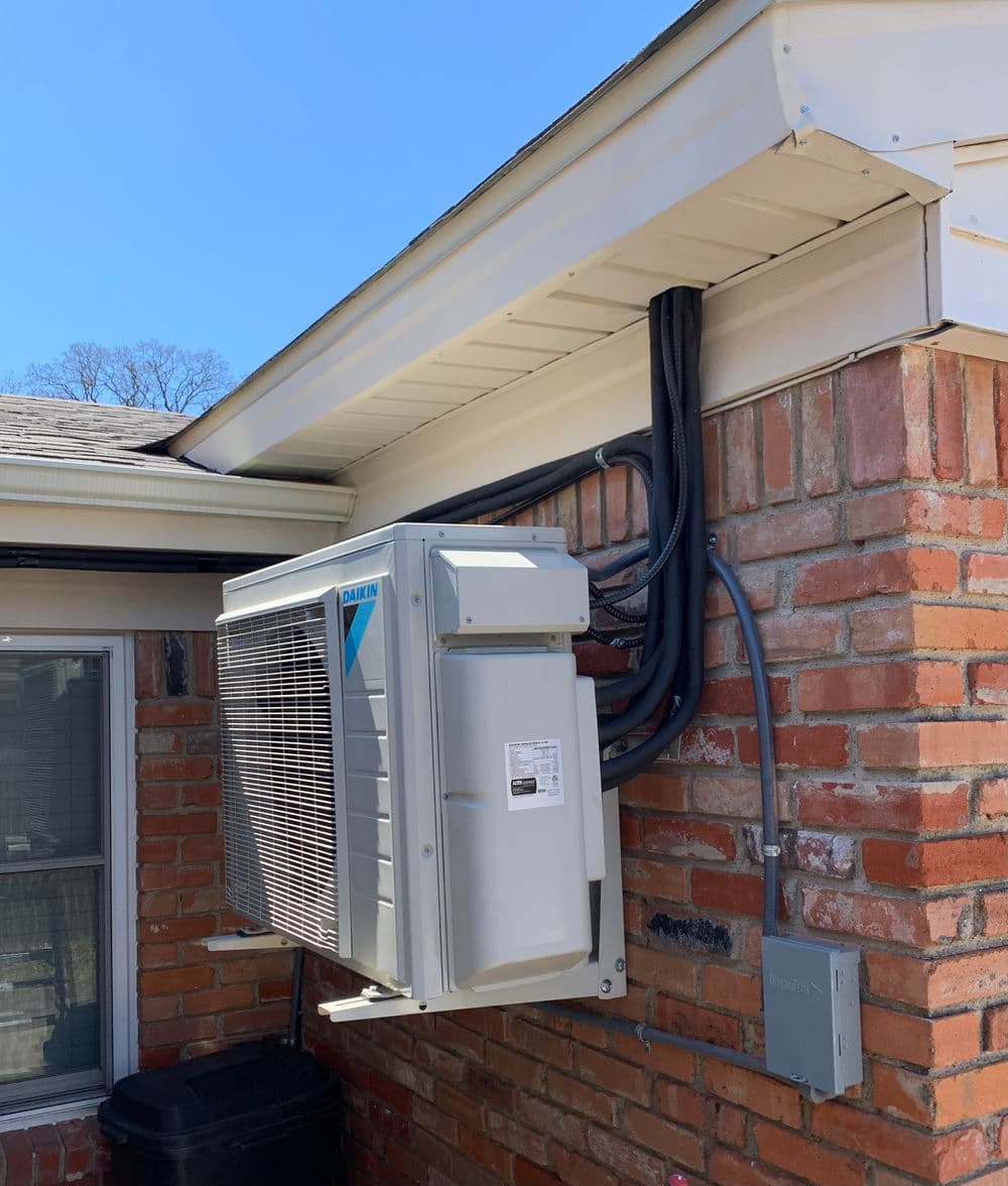 Outdoor air conditioning unit mounted on a brick wall with connecting pipes and electrical box.
